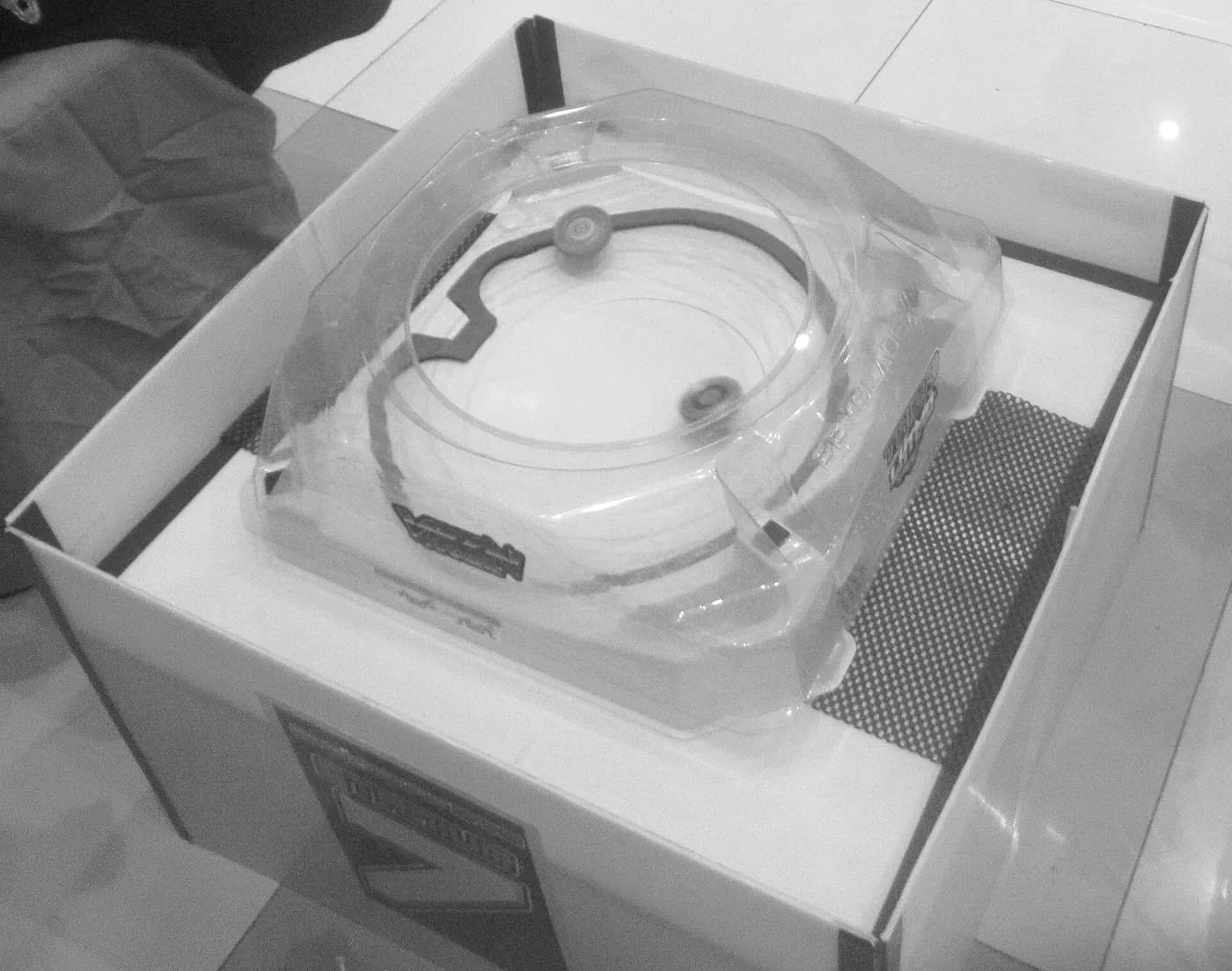Un'arena trasparente di Beyblade X dentro una scatola, con due trottole che girano sul bordo